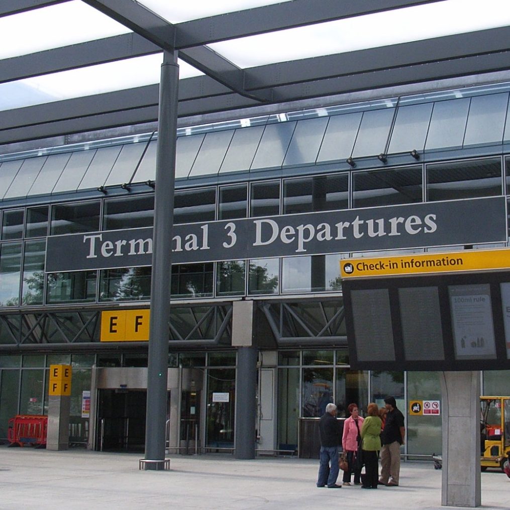 A person wearing a mask walks through Heathrow Airport, pulling a suitcase.