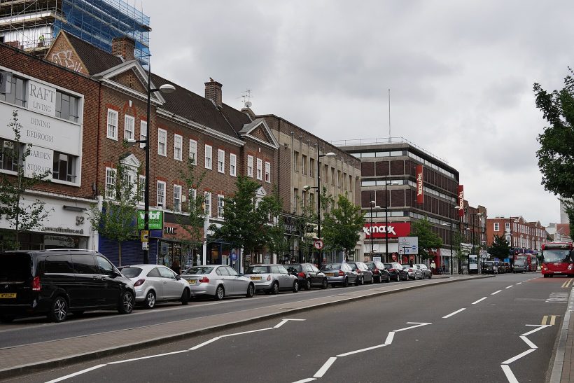 Street view featuring shops and parked cars along a side road under a cloudy sky.