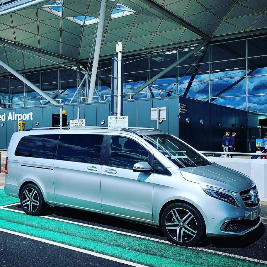A silver six-seater car is parked at an airport, with an airport building in the background.