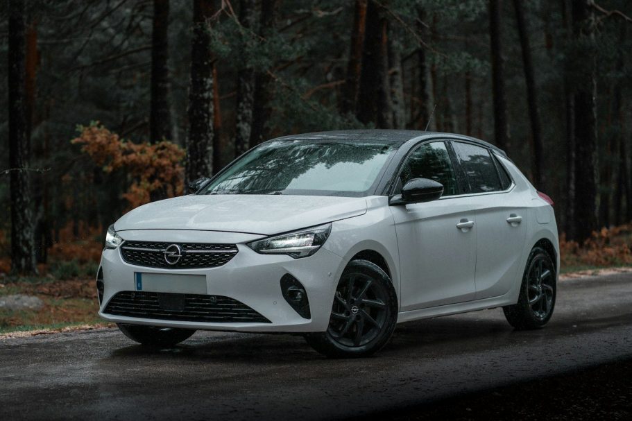 White hatchback car parked on a forest road surrounded by trees.