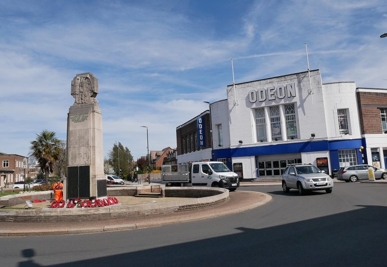 A war memorial surrounded by flowers, with a building and vehicles nearby.